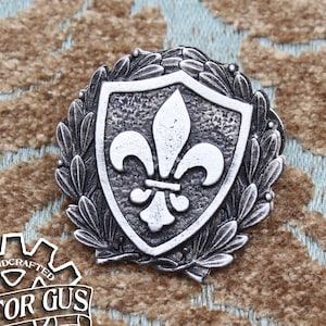 May include: A silver-toned metal pin featuring a shield with a fleur-de-lis design. The shield is surrounded by a wreath of leaves. The pin is handcrafted and has the text "Doctor Gus Original" on the back.
