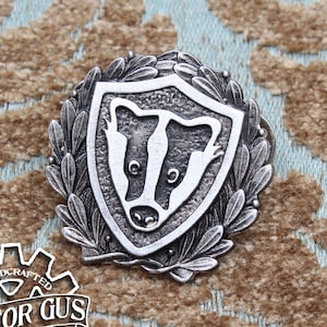 Può includere: Uno spillo in metallo argentato con una testa di tasso all'interno di uno scudo circondato da una corona di alloro. Lo spillo è fatto a mano e dice "Doctor Gus Original" sul retro.