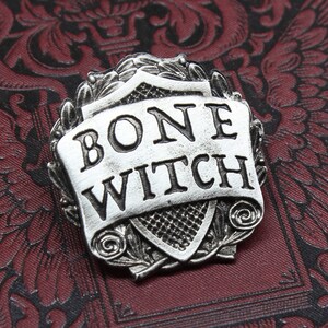 May include: A silver-toned metal pin with a shield design. The shield has a scroll with the words "BONE WITCH" on it. The pin is handcrafted by Doctor Gus.