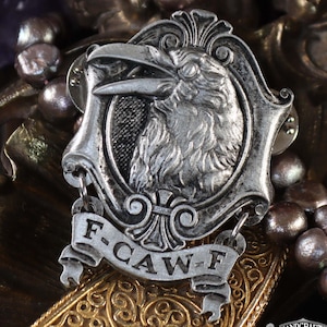 May include: A silver-toned raven pin with the text "F-CAW-F" on a banner. The raven is inside a decorative frame. The pin is set against a backdrop of ornate gold and purple elements, with the Doctor Gus logo in the bottom right corner.