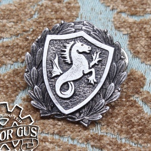 May include: A silver-toned metal pin featuring a stylized seahorse within a shield shape, surrounded by a wreath of leaves. The pin is handcrafted and has the text "Doctor Gus Original" on the back.