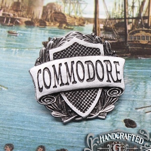May include: A silver-toned metal pin with a shield design and the word "COMMODORE" on a banner. The pin is surrounded by a wreath of leaves. The pin is handcrafted by Doctor Gus.