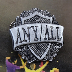 May include: A silver-toned pin with a shield design and the words "ANY/ALL" on a banner. The pin is handcrafted and features a detailed design.