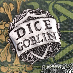 Op de afbeelding: Een zilverkleurige metalen pin met een scroll-ontwerp en de woorden "Dice Goblin" erop. De pin is handgemaakt door Doctor Gus.