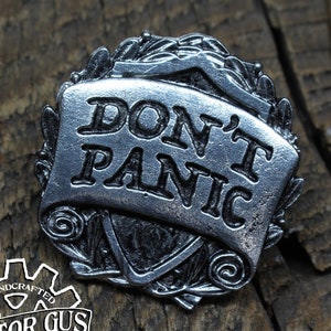 May include: A silver-toned metal pin with a scroll design that reads "Don't Panic". The pin is surrounded by a wreath of leaves and is handcrafted by Doctor Gus.