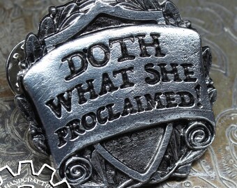 May include: A silver-toned metal pin with a scroll design and the text "Doth What She Proclaimed!" The pin is on a textured, metallic surface.