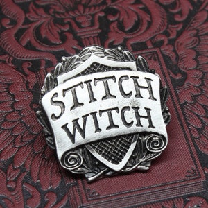 May include: A silver-colored metal pin with the words "STITCH WITCH" on a banner. The pin is set against a dark red background with an ornate floral pattern. The pin has a shield-like shape with decorative elements.