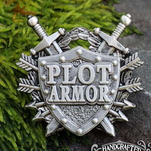 May include: A silver-colored metal pin featuring a shield with the words "PLOT ARMOR" in bold letters. The shield is surrounded by crossed swords, arrows, and laurel leaves. The pin is set against a backdrop of green moss and a stone surface.