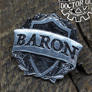 May include: A silver-toned metal pin with a shield design and the word "BARON" in black lettering on a scroll. The pin is handcrafted by Doctor Gus.
