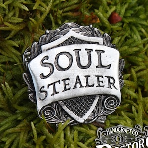 May include: Silver-toned metal pin with a shield design and the words "Soul Stealer" on a scroll. The pin is handcrafted by Doctor Gus Original.