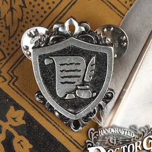 May include: A silver charm in the shape of a shield with a scroll and quill pen inside. The charm is on a brown background with a feather.