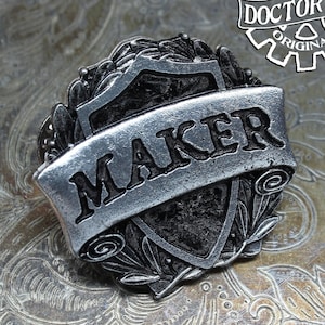 May include: A silver-toned metal pin with the word "MAKER" on a banner, set against a shield and laurel wreath design. The pin is stamped with "HANDCRAFTED DOCTOR GUS ORIGINAL" in a gear-shaped logo. The background is a textured, metallic surface.
