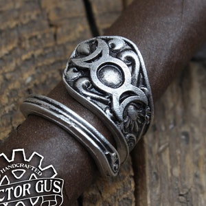 May include: A silver ring with a detailed design featuring a circle with three crescent moons and floral accents. The ring is handcrafted and has the text "Doctor Gus Original" on the side.