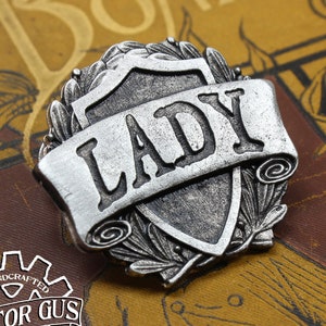 May include: Silver-toned metal pin with a shield design and a scroll banner that reads "LADY". The shield is surrounded by a wreath of leaves.