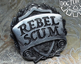 May include: A silver-toned metal pin with a shield-shaped design. The shield has a banner that reads "Rebel Scum". The pin is handcrafted by Doctor Gus Original.