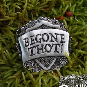May include: A silver-toned metal pin with a scroll design and the words "BEGONE THOT!" in raised lettering. The pin is on a bed of green moss.