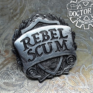 May include: A silver-toned metal pin with a shield-shaped design. The shield has a banner that reads "Rebel Scum". The pin is handcrafted by Doctor Gus Original.