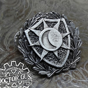 May include: A silver-toned metal pin featuring a shield with a sun and moon design. The shield is surrounded by a wreath of leaves. The pin is handcrafted and has the text "DOCTOR GUS ORIGINAL" on the back.