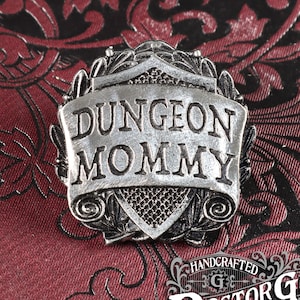 May include: A silver-toned pin with a scroll design and the text "Dungeon Mommy". The pin is handcrafted by Doctor Gus Original.