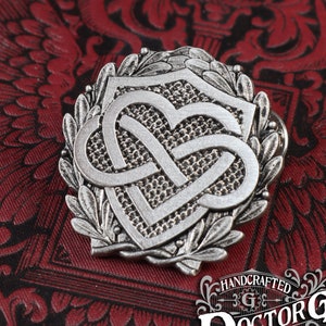 May include: A silver-toned metal pin with a Celtic knot design in the shape of a heart. The knot is surrounded by a wreath of leaves and is set against a red background. The pin is handcrafted and has the text "DoctorGus Original" on the bottom.
