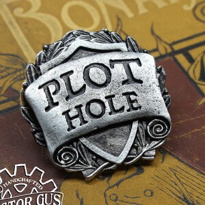 May include: A silver-toned metal pin with a shield-shaped design. The shield has the words "PLOT HOLE" written on it in a scroll-like banner. The pin is handcrafted by Doctor Gus.