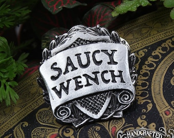 May include: A silver-toned metal pin with a scroll design and the words "Saucy Wench" on it. The pin is on a black and gold patterned surface.