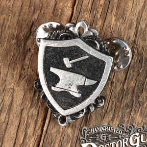 Könnte beinhalten: Eine silberfarbene Metallstifte mit einem schwarzen Schild mit einem stilisierten Amboss und Hammer. Das Schild ist von dekorativen Schnörkeln umgeben. Der Stift ist handgefertigt von Doctor Gus Original.