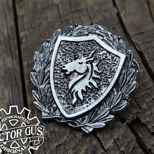 May include: A silver-toned metal pin featuring a shield with a stylized horse head design. The shield is surrounded by a wreath of leaves. The pin is handcrafted and has a textured surface.