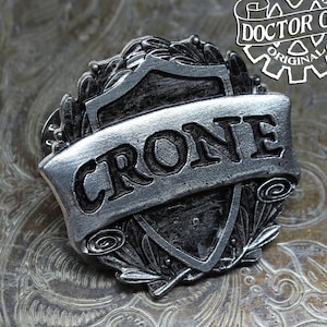 May include: A silver-toned metal pin with a shield design. The shield has a black background and a silver-toned border. The word "CRONE" is written in black letters on a silver-toned banner across the shield. The pin is decorated with silver-toned leaves.