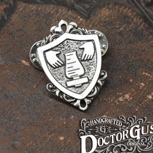 May include: Silver-toned metal lapel pin with a shield design. The shield features a stylized illustration of hands and a measuring device. The pin is adorned with decorative flourishes and the text "HANDCRAFTED DOCTOR GUS ORIGINAL".