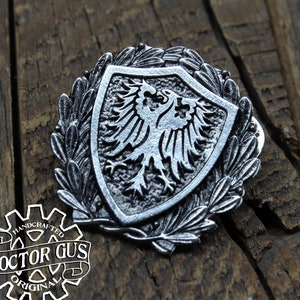 May include: A silver-toned metal pin featuring a shield with a double-headed eagle design. The shield is surrounded by a wreath of leaves. The pin is handcrafted and has the text "DOCTOR GUS ORIGINAL" on the back.