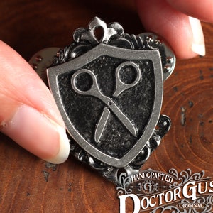 May include: Silver-toned metal pin featuring a shield with a pair of scissors inside. The shield is surrounded by an ornate design. The pin is handcrafted by Doctor Gus.