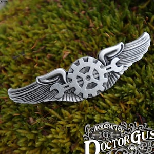 May include: Silver-toned metal pin with a gear and wrench design, set against a green mossy background. The pin features a pair of wings and the text "HANDCRAFTED DOCTOR GUS ORIGINAL".