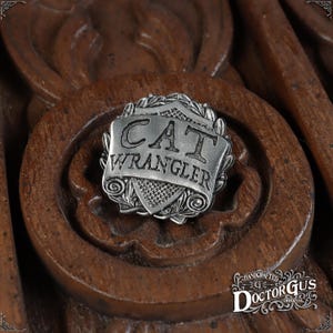 Puede incluir: Un pin de metal plateado con las palabras "CAT WRANGLER" en un rollo. El pin está hecho a mano por Doctor Gus Original.