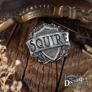 May include: A silver-toned metal pin featuring a shield design and the word "SQUIRE" on a banner. The pin is handcrafted by Doctor Gus Original.