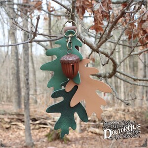 May include: A leather keychain featuring two oak leaves, one green and one brown, with a small acorn-shaped bell. The keychain is hanging from a tree branch. The text "HANDCRAFTED DOCTOR GUS ORIGINAL" is visible in the bottom right corner of the image.