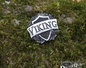May include: A silver-toned metal pin with a shield design and the word "VIKING" on a banner. The pin is surrounded by a wreath of leaves.