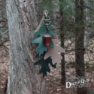 May include: A leather acorn charm with a green and brown oak leaf design. The charm is attached to a silver key ring and hangs from a tree branch. The charm is handcrafted by Doctor Gus Original.