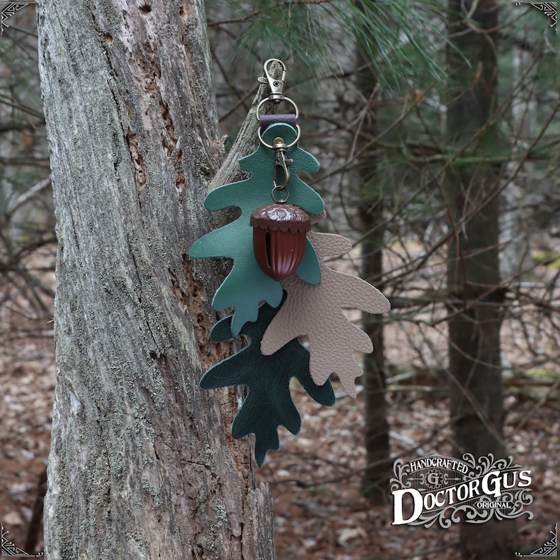 May include: A leather acorn charm with a green and brown oak leaf design. The charm is attached to a silver key ring and hangs from a tree branch. The charm is handcrafted by Doctor Gus Original.