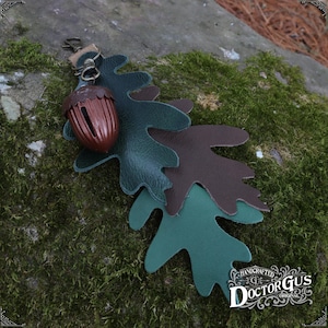 May include: A leather acorn charm with three layered oak leaves in shades of green and brown. The charm is attached to a metal ring and has the text "HANDCRAFTED DOCTORGUS ORIGINAL" on the bottom.