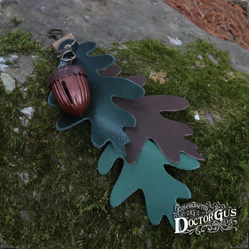 May include: A leather acorn charm with three layered oak leaves in shades of green and brown. The charm is attached to a metal ring and has the text "HANDCRAFTED DOCTORGUS ORIGINAL" on the bottom.