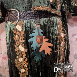 May include: A green velvet dress with gold embroidery and a brown leather belt. A green and brown leather acorn charm hangs from the belt.