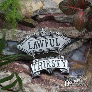 May include: A silver-toned metal pin with two hanging signs. The top sign reads "LAWFUL" and the bottom sign reads "THIRSTY". The pin is handcrafted by Doctor Gus Original.