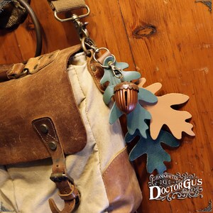 May include: A brown leather bag with a canvas strap and a leather acorn charm with two green and brown leather leaves attached. The charm is hanging from a metal key ring. The bag has a metal buckle and a brown leather flap. The text "HANDCRAFTED DOCTOR GUS ORIGINAL" is visible in the lower right corner of the image.