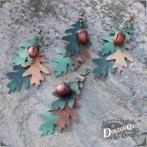 May include: Three leather oak leaf keyrings with acorn charms. Each keyring features three layered leaves in shades of green, brown, and tan. The acorns are a dark brown colour with a gold-toned top.