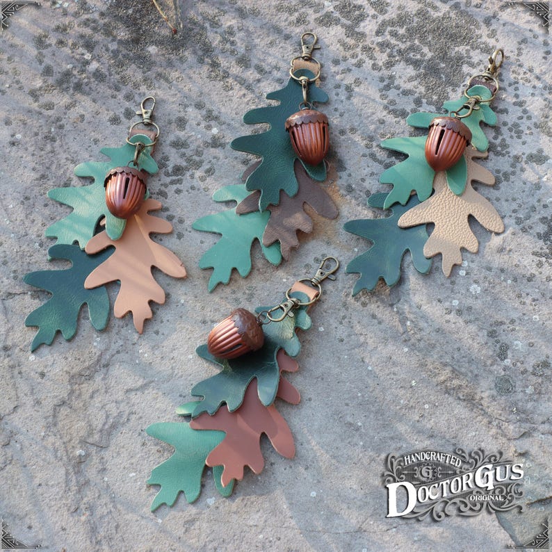 May include: Three leather oak leaf keyrings with acorn charms. Each keyring features three layered leaves in shades of green, brown, and tan. The acorns are a dark brown colour with a gold-toned top.