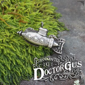 Könnte beinhalten: Eine detaillierte Miniatur-U-Boot-Anstecknadel in Silber mit Steampunk-Ästhetik. Die Anstecknadel verfügt über einen Propeller, Bullaugen und Nieten. Die Anstecknadel ist handgefertigt von Doctor Gus Original.