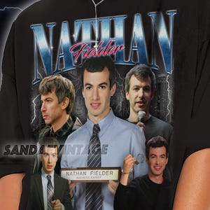 Pode incluir: T-shirt preto com um gráfico de Nathan Fielder e o texto "NATHAN Fielder" em azul e rosa. O design inclui várias imagens de Nathan Fielder. A camisa é de estilo vintage.