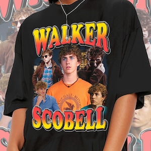 Op de afbeelding: Zwart t-shirt met de naam "WALKER SCOBELL" in retrostijl. De shirt toont afbeeldingen van een jonge man in verschillende poses. "WALKER" is in een gele en rode gradiënt, en "SCOBELL" is in geel en rood.