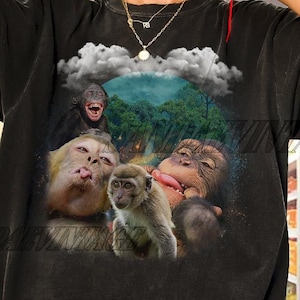 May include: Black t-shirt featuring a graphic of several monkeys with their tongues out, set against a backdrop of a cloudy sky and lush green foliage. The shirt has a vintage, worn look.